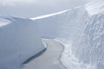 A winding road cuts through towering snowdrifts, creating a serene and isolated winter landscape under a bright sky.