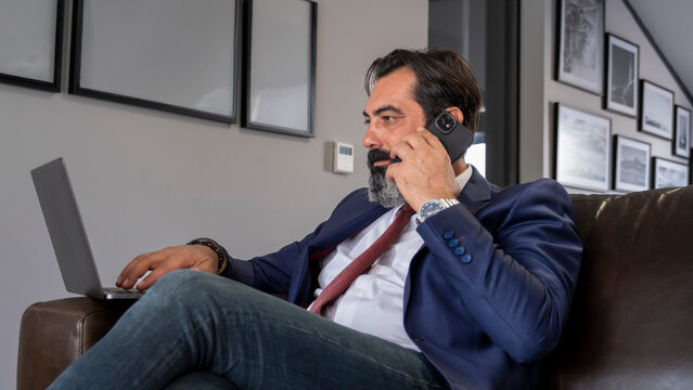 A middle-aged, dark-haired businessman in a suit is sitting on a leather couch in his office, having a tense business meeting on his phone