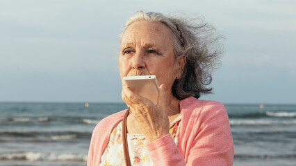 Senior woman with grey hair sending a voice message on her smartphone at the beach