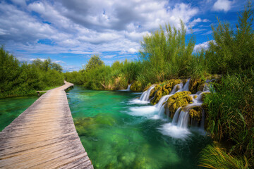 A scenic wooden walkway follows a clear turquoise river with small waterfalls cascading over mossy rocks, surrounded by lush green trees under a bright blue sky with clouds