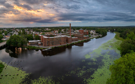 Aerial view of a brick building complex with a tall chimney reflecting in the still river, contrasted by the green algae and the dramatic sky, Waltham, Middlesex County, United States.