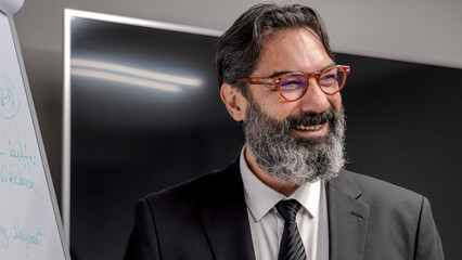 A middle-aged, dark-haired businessman is in a conference room, standing in front of a whiteboard and explaining something