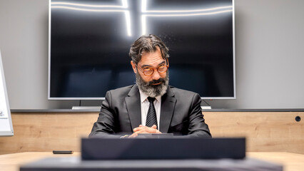 A middle-aged, dark-haired, bearded businessman in a suit is sitting at a conference table in his office, looking at his employees with a tense expression during a meeting