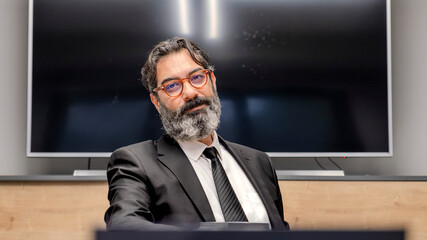 A middle-aged, dark-haired businessman in a suit is happily working at his desk in the office smiling as he looks at the camera