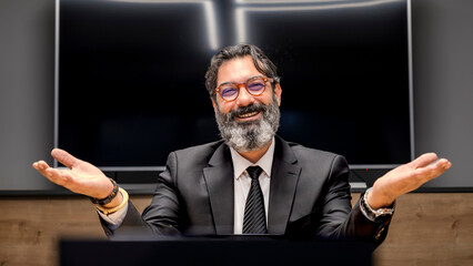 A middle-aged, dark-haired businessman in a suit is happily working at his desk in the office smiling as he looks at the camera