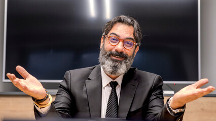 A middle-aged, dark-haired businessman in a suit is happily working at his desk in the office smiling as he looks at the camera
