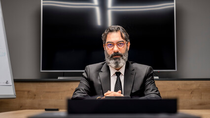 A middle-aged businessman with dark hair, dressed in a suit, is sitting at his desk in the office,...