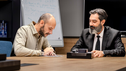 One dark-haired, bearded middle-aged businessman and another businessman are sitting around a table in a conference room, enjoying a pleasant business meeting in an office environment