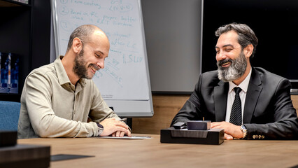 One dark-haired, bearded middle-aged businessman and another businessman are sitting around a table in a conference room, enjoying a pleasant business meeting in an office environment