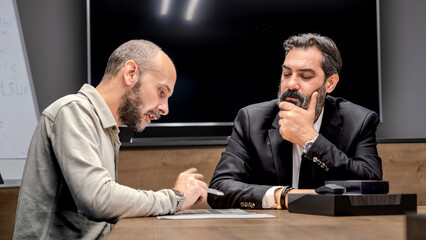One dark-haired, bearded middle-aged businessman and another businessman are sitting around a table in a conference room, enjoying a pleasant business meeting in an office environment