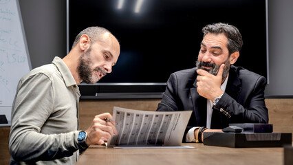One dark-haired, bearded middle-aged businessman and another businessman are sitting around a table in a conference room, enjoying a pleasant business meeting in an office environment