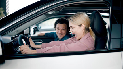 Young woman excitedly test drives a new car in a showroom, while a smiling man observes, showcasing...