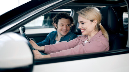 Young woman is joyfully testing car features while man observes in a modern car showroom,...