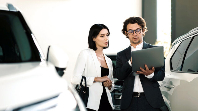A woman discusses car options with a salesperson in a modern showroom where various car models are displayed and a laptop is used for information.