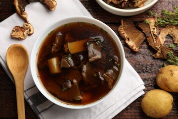 Tasty soup in bowl, raw potato and dry chanterelle mushrooms on wooden table, flat lay