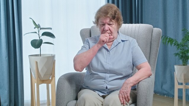 Mature woman in armchair feeling unhealthy and coughing, flu symptoms and treatment concept