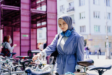 Muslim woman in lilac blazer unlocking bicycle near pink bike station