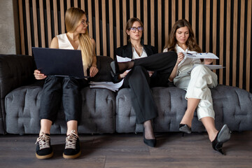 Businesswomen reviewing project data on paper and laptop while seated on office couch