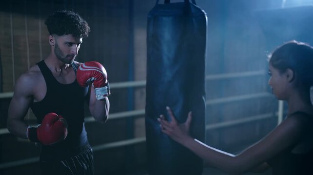 Indian young adult gen z angry fit boxer man guy wear gloves do hand fist learn boxing punch beat on bag indoor dark room place modern sports female girl teach to male self defense martial art skill