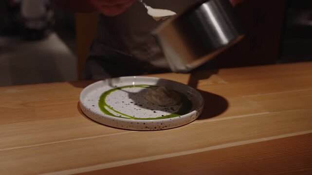Unrecognized skilled chef carefully pours vibrant green sauce onto plate, showcasing culinary artistry. The upscale kitchen setting enhances the elegant presentation of the dish.