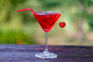 Bright red cherry cocktail in a martini glass against summer garden background on wooden table, closeup. Delicious cold alcoholic drink for relaxation on a sunny warm day
