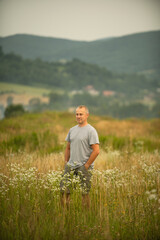 A man stands in a field of tall grass, holding a microphone as if he is about to speak or sing. The...