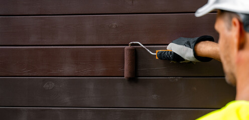 house painter with paint roller painting wooden exterior siding with protective color. wood...