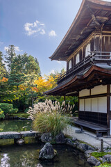 Ginkaku-ji temple building in Kyoto, Japan, seen from garden pond with reflection, framed by seasonal plants, pine trees, and curved tiled roof in vertical composition