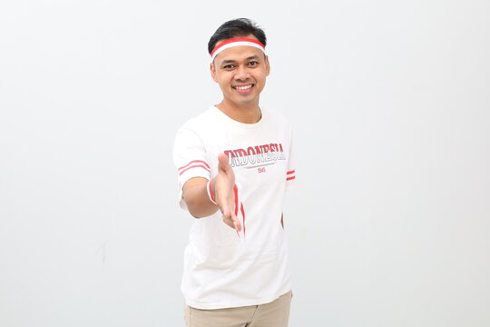 Indonesian man wearing red and white ribbon celebrating Indonesian independence day smiling while standing extending hand in handshake gesture looking at camera, isolated on white background.