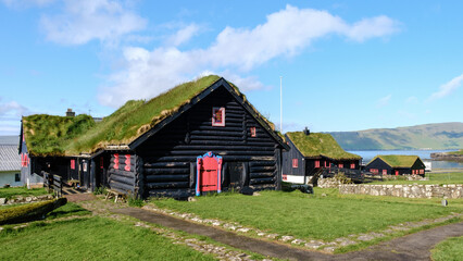 Quaint black wooden homes with grass roofs dot the lush green landscape of the Faroe Islands....