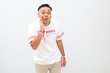 Naklejka premium Indonesian man with red and white ribbon celebrating Indonesian independence day, standing looking at camera with hand beside mouth, gesture of making announcement or whispering secret.