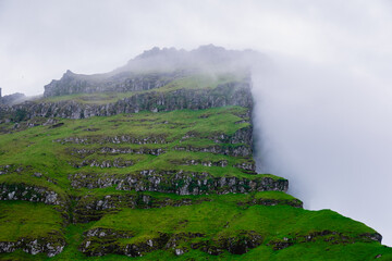 Rugged cliffs tower over verdant landscapes in the Faroe Islands, enveloped by mist and clouds. The...