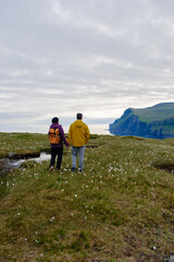 Obraz premium A couple strolls hand in hand through lush green meadows adorned with delicate flowers in the Faroe Islands. The dramatic cliffs and serene ocean in the background create a breathtaking backdrop.