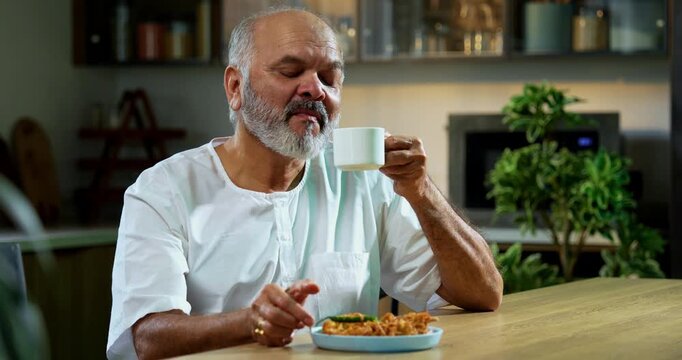 Senior Indian man eating pakoda or pakora with tea on table, Asian foodie old man expressing immense happiness while enjoying tasty onion fritters in modern kitchen setting, joyful retired person