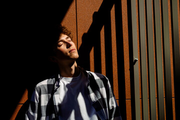 A young man with closed eyes stands by the wall, illuminated by the sun's rays. His shadow falls on...