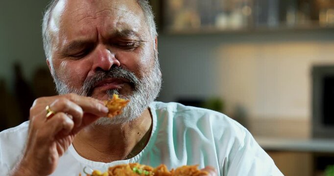 Senior Indian man eating pakoda or pakora with tea on table, Asian foodie old man expressing immense happiness while enjoying tasty onion fritters in modern kitchen setting, joyful retired person