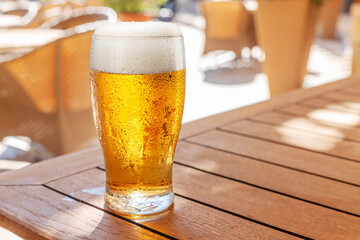 Chilled glass of light beer sweating in the sun on wooden table and blurred summer terrace at the background.