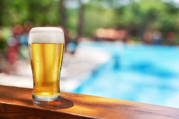 Glass of chilled beer on table and blurred sparkling pool water at the background. Place for your product or brand name display.