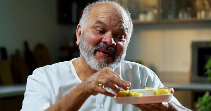 Senior Indian man eating khaman dhokla with joyful expression, Asian foodie old man enjoying tasty Gujarati snack while sitting at table in modern kitchen, expressing immense happiness and delight