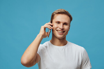 Smiling man talking on mobile phone with friendly expression against blue background, casual white...