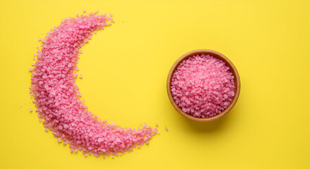 Pink Himalayan bath salt crystals arranged in a crescent moon shape next to a small wooden bowl on a yellow background, perfect for spa and wellness.
