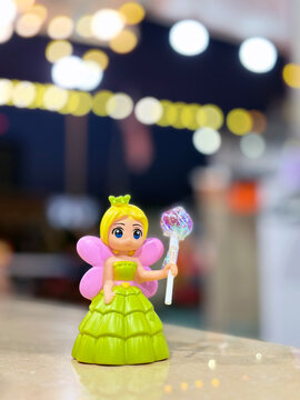 A small, vibrant fairy doll in a green dress with pink wings and a shimmering wand stands on a surface, with blurred, festive bokeh lights in the background.