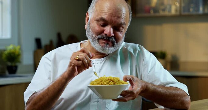 Senior Indian man eating noodles joyfully at home in modern kitchen, Asian foodie old man expressing immense happiness, satisfaction while enjoying tasty snack at dining table in bright home setting