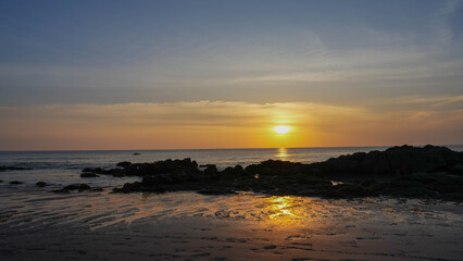 Fototapeta premium Title: Orange sunlight reflects off the calm ocean as the sun sets over the horizon, painting the sky with beautiful clouds