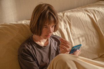 Teenage boy engaged with smartphone while sitting on couch at home during afternoon light
