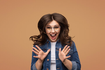 Excited young woman with glasses in a denim jacket, gesturing energetically and smiling brightly....