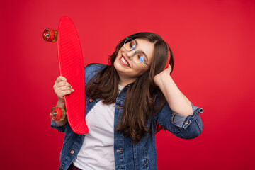 Young woman in glasses and denim jacket holding a red skateboard while smiling playfully.