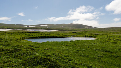 bering island. Lush green landscape with a serene pond surrounded by rolling hills and patches of snow under a bright blue sky, showcasing the beauty of nature and tranquility