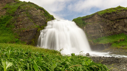 bering island. Majestic waterfall cascading over rocky cliffs surrounded by lush green grass and...