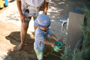 Child Filling Bucket with Water from Outdoor Tap Under Adult Supervision